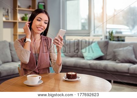 Coffeebreak in cozy cafe. Beautiful brunette with smartwatch having cup of coffee and dessert. Nice coffee house interior. Big window as a background. Woman using mobile phone and smiling