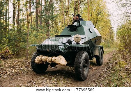 Pribor, Belarus - April 24, 2016: Re-enactor Dressed As Russian Soviet Crew Member Soldier Of World War II Sitting In Armoured Soviet Scout Car BA-64 In Forest And Aiming a Sub-machine Gun at Enemy