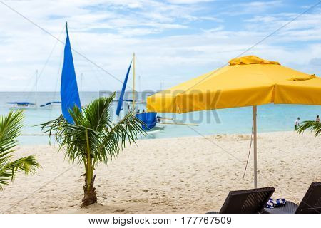 Boracay Beach small palm trees and a yellow parasol in the foreground of a white sand beach with calm sea and Filipino sailing pump boats just off shore.