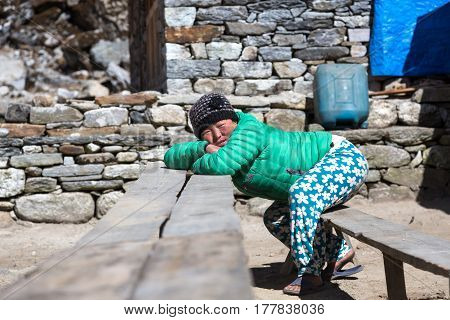 Portrait of Nepalese Girl in traditional colorful Pants and Down Vest relaxing on wooden Bench leaning on Table. Kothey Lodge, Nepal, Solo-Khumbu region, November 3, 2016
