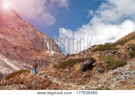 Sunny Mountains View rocky Trail and Hiker in red Jacket and blue Pants walking up carrying backpack and using trekking poles