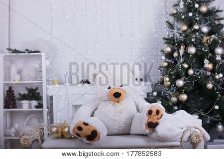 Big white fluffy teddy bear sitting on the bench in white beautiful Christmas studio. Christmas room interior decorations in studio.