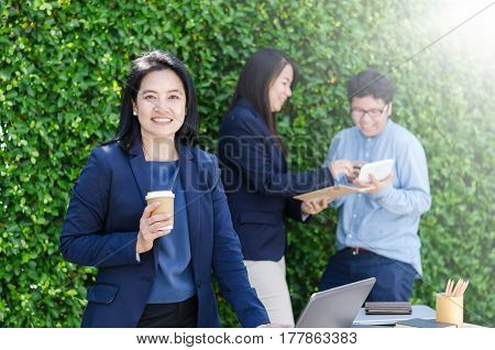 Smiling Asian Woman Holding Cup Of Coffee And Her Team Work Dicussing.
