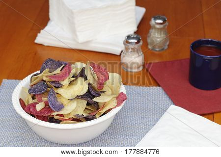 One bowl of red white and blue potato chips on a kitchen table with linens seasonings and beverage. Red white and blue potato chips in a bowl on a kitchen table.