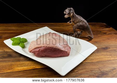 Dinosaur Tyrannosaurus Rex Staring A Full Piece Of Raw Cow Beef (picanha), With Basil On A White Dis