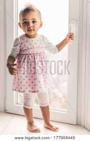 Cute Afro American baby girl is looking at camera while standing on the window sill at home
