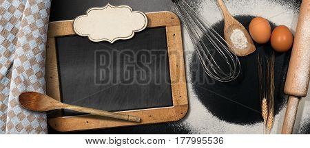 Baking background with empty blackboard and label on a black table with rolling pin whisk eggs spoons ears of wheat and a checkered tablecloth