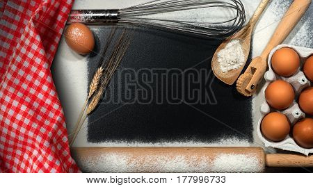 Baking background - Kitchen utensils ears of wheat eggs checkered tablecloth and flour with rectangular copy space on a black table