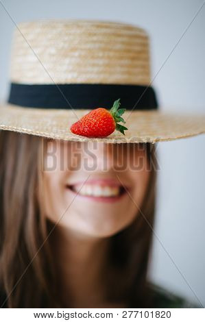 Portrait Of Brunette Girl In Straw Panama Hat With Strawberry On It Against White Wall Background. F