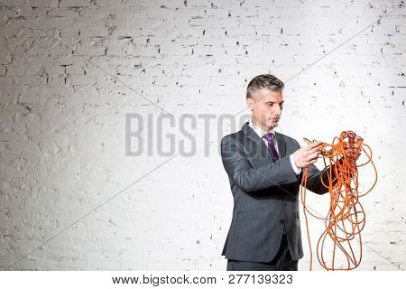 Confident mature businessman holding tangled red cable against white brick wall