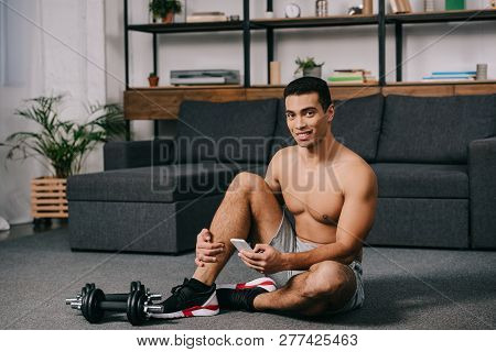 Cheerful Bi-racial Man Sitting On Floor Near Dumbbells While Using Smartphone In Living Room