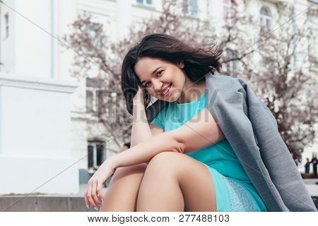 Beauitful plus size model wearing blue dress and grey coat walking by blooming spring tree outdoor on the city street.
