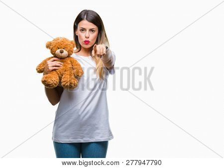 Young beautiful woman holding teddy bear plush over isolated background pointing with finger to the camera and to you, hand sign, positive and confident gesture from the front
