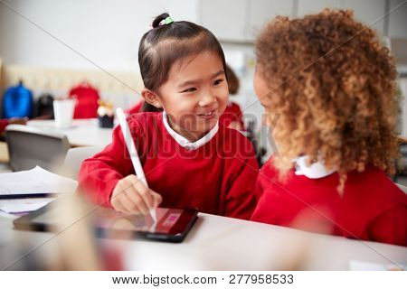 Close up of two kindergarten schoolgirls wearing school uniforms, sitting at a desk in a classroom using a tablet computer and stylus, looking at each other smiling