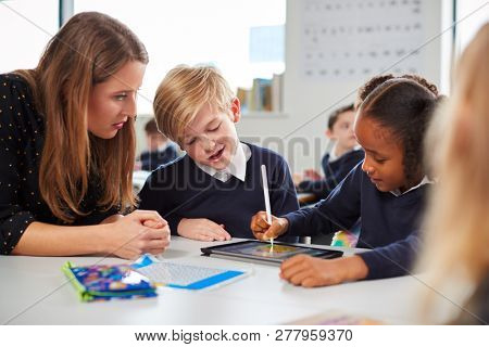 Female school teacher helping two kids using a tablet computer at desk in a primary school classroom, close up