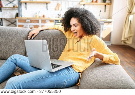 Unexpected News. Surprised Multi-ethnic Girl Lies On A Comfortable Sofa With A Laptop And Looks At T