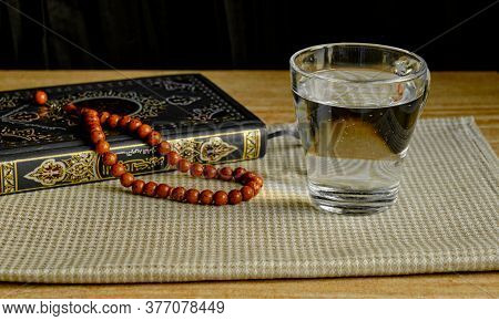 Glass Of Clear Water, Stone Rosary And Green And Gilded Book Of Quran On Napkin During Sunset. First