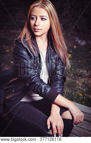 Calm Blonde Is Sitting On Parkbench In Black Jacket And Looking Away Of Camera