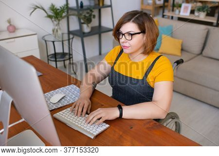 Young pretty female in casualwear sitting in wheelchair by desk and looking at computer screen while learning remotely from home