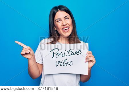Young beautiful woman asking for optimist attitude holding paper with positive vibes message smiling happy pointing with hand and finger to the side