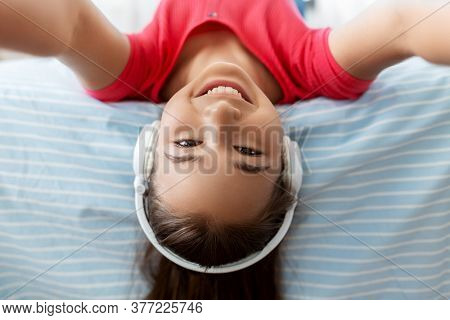 children and technology concept - portrait of smiling teenage girl in headphones lying on bed upside down and listening to music and taking selfie at home