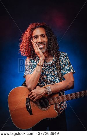 Talanted Hispanic Musician In A Hawaiian Shirt Holds Guitar, Smiling And Looking On Camera. Studio S