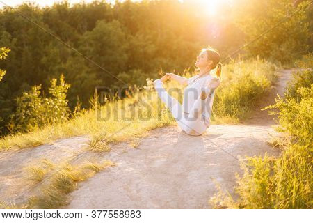 Young Fit Woman Performing Yoga Posture Boat With Toe Hold, Outside Evening On Background Of Bright 