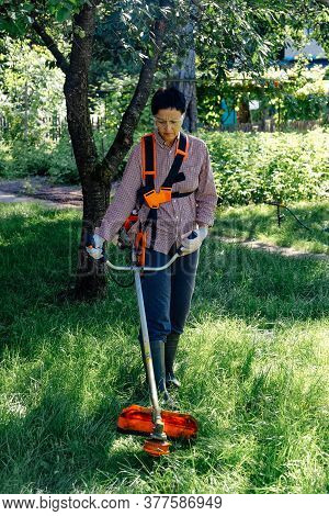 Female Worker Mowing Lawn With Grass Trimmer Outdoors In Garden.