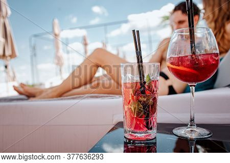 Young Hot Couple Resting At Swimpool. Two Red Cold Tasty Cocktails Stand On Table. Man And Woman Lyi