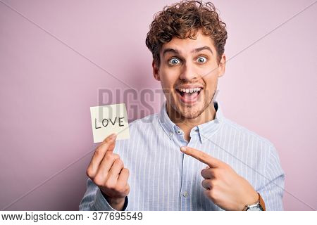 Young blond romantic man with curly hair holdiing paper with love message for valentines day very happy pointing with hand and finger