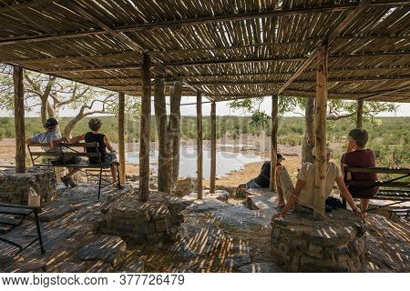 Halali, Namibia - April 3, 2019 : Tourists Wait For Wildlife Sitting On A Bench At The Moringa Water