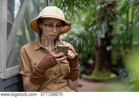 Woman Botanist Dressed In Safari Style In Greenhouse. Naturalist In Khaki Clothes, Hat, Leaver Glove