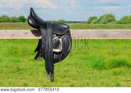 The Black Saddle And Its Girth Is Lying On The Wooden Hitching Post In Outdoors.