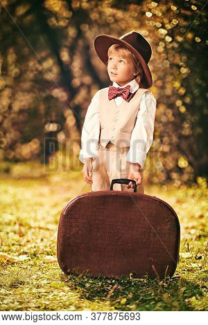 Happy little boyboy in a classic suit and hat possing with a large brown retro suitcase in a beautiful autumn park. Retro style. Children's fashion.
