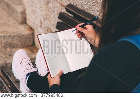 Young Woman Sits On Bench In Park With Blank Notebook And Pen On Her Hands. Wish Marathon. Writing W