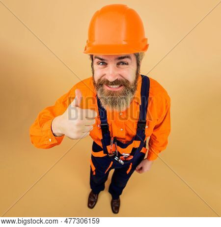 Smiling Bearded Man In Overalls And Hardhat Showing Thumb Up. Builder In Safety Helmet And Uniform I