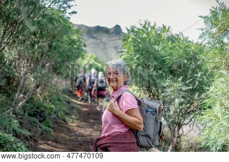 Carefree Caucasian Senior Woman Carrying Backpack Looks At Camera Walking The Trail On A Mountain Hi