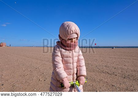 Little Girl In A Coat Dragging Her Scooter Through The Sand On An Empty Beach