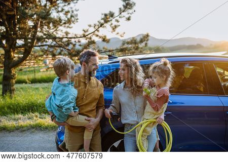 Happy Family In Front Of Their Electric Car, Concept Of Green Energy And Sustainable Lifestyle.