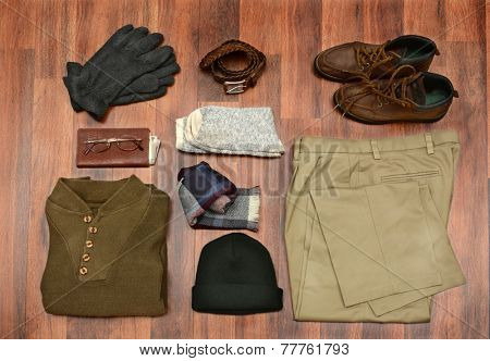High angle shot of men's winter clothes laid out on a dark wood floor. Items include, Sweater, Scarf, Gloves, Wool Socks, Pants, Boots, Belt, Knit Cap, Wallet, and Glasses. Horizontal Format.