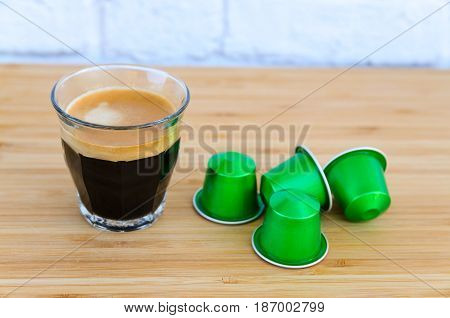 Close up of tasty espresso with foam and green coffee capsules on wooden background.