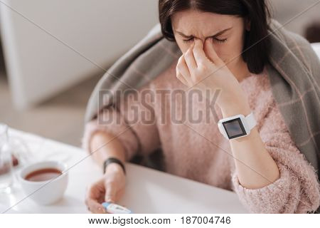 Hope for improvement. Photo of ill female person wearing fluffy sweater keeping eyes closed while holding thermometer