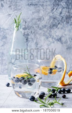 Tonic water cocktail with rosemary, blueberries and orange. Two cold glasses and bottle with zest and bubbles over gray blue texture background. Refreshing beverage alco non alcohol