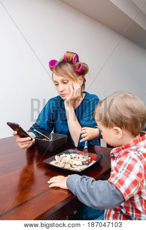 Bored tired white Caucasian young woman mother housewife with hair-curlers in hair looking on phone surfing Internet f child son boy sitting eating meal lunch lifestyle concept