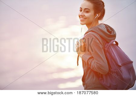 Portrait of beautiful happy girl in sportswear with small urban backpack on the shore of a lake or sea