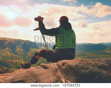 Tired Tourist With Medicine Crutch  And Broken Leg Fixed In Immobilizer Resting On Summit.