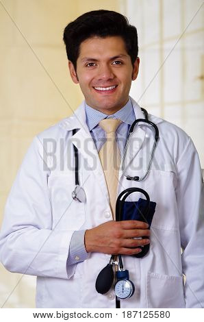 Confident handsome smiling doctor posing and looking at camera with an Stethoscope around his neck and holding a tensiometer in his hand, in office background.