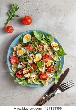 Farfalle bow tie pasta salad with quail eggs, cherry tomatoes, rucola, mozzarella and basil, dressed with balsamic sauce over grey concrete background