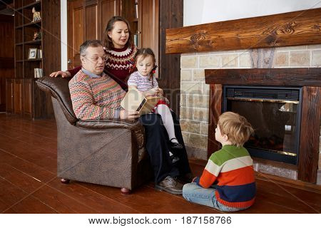 Father reads book aloud sitting in armchair near fireplace, mother and two children liten to him.