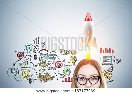 Close up of an Asian woman s head in glasses near a gray wall with a colorful rocket start up sketch